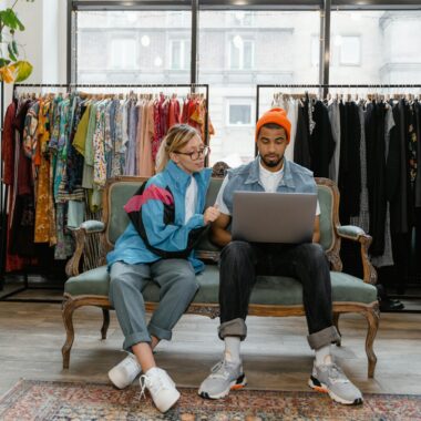 A couple sits on a couch in a stylish clothing store, browsing a laptop together.